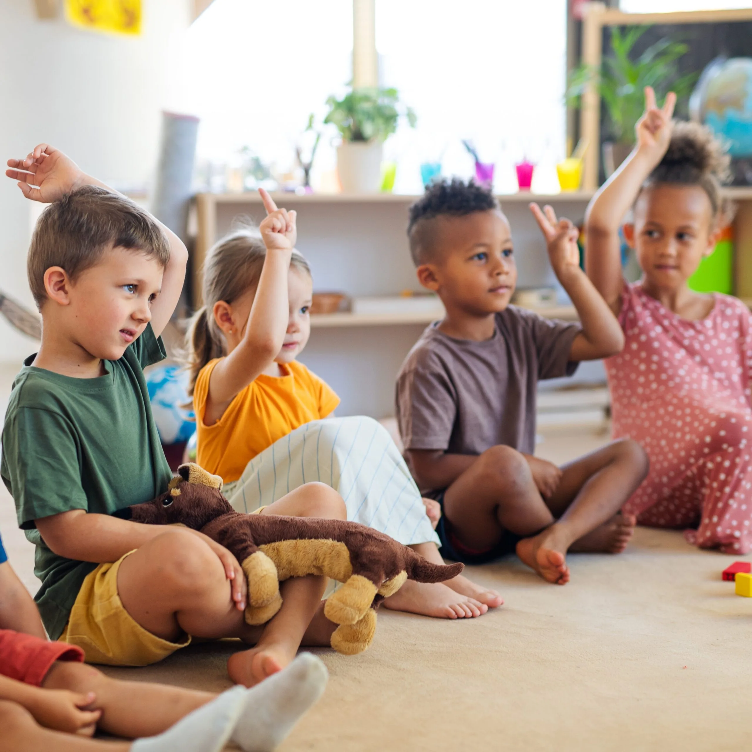A group of small nursery school children sitting on floor indoors in classroom, raising hands.
